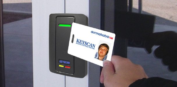 Verkada Installers A person holds a Keyscan access card up to an electronic card reader to unlock a glass door, showcasing Jendy Physical Security’s commitment to advanced access control. The card displays a photo ID and text.