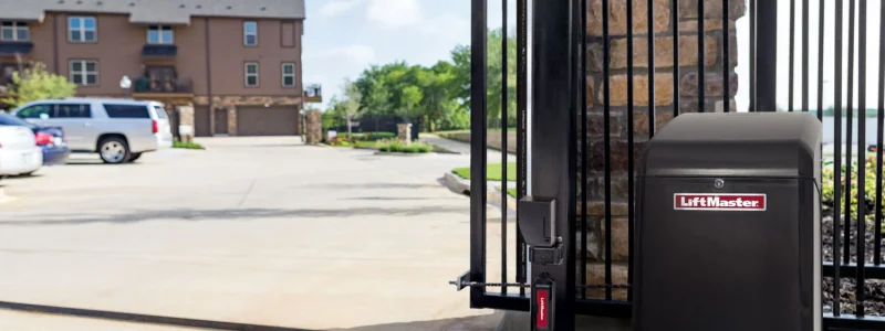 Verkada Installers Black LiftMaster gate operator installed on a metal gate at the entrance to a residential complex, with an apartment building and parked vehicles visible in the background by Jendy Physical Security.